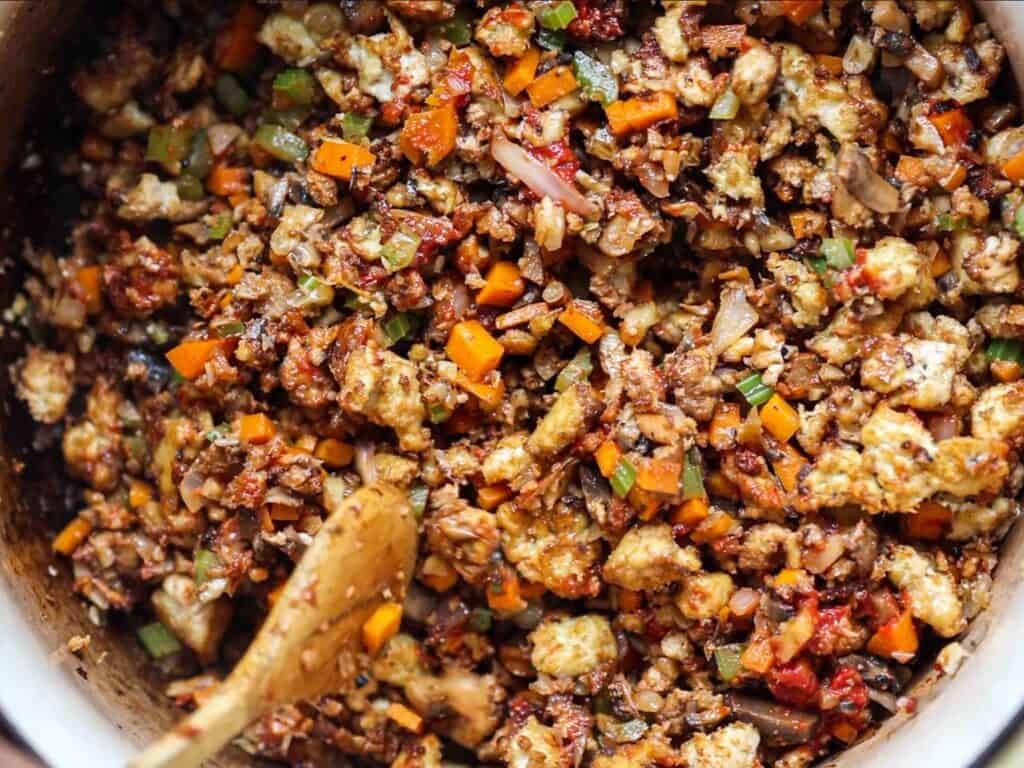 A close-up of a colorful, mixed stuffing with diced vegetables, tofu, and seasonings. A wooden spoon is partially visible, stirring the mixture in a pot.